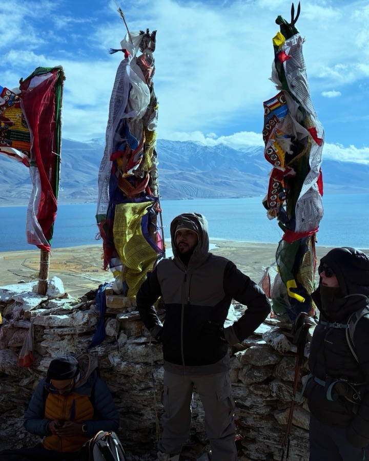 Pratik standing among prayer flags overlooking Tso Moriri lake in Ladakh, snow peaks in the distance